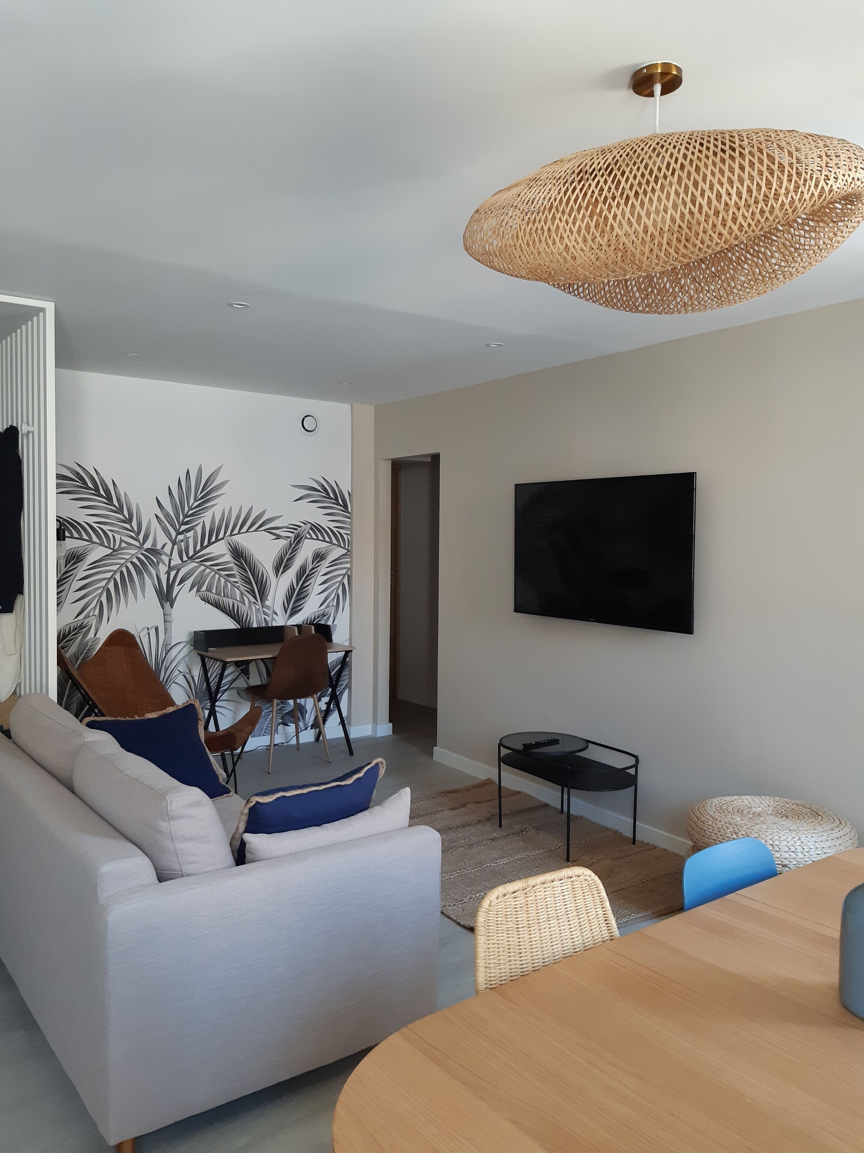 A modern living room with a sofa, dining table, and chairs. There is a flat-screen TV mounted on the wall and a woven pendant light fixture.
