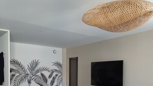 Een moderne woonkamer met een bank, eettafel en stoelen. Er is een flatscreen televisie aan de muur bevestigd en een geweven hanglamp.