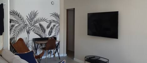 A modern living room with a sofa, dining table, and chairs. There is a flat-screen TV mounted on the wall and a woven pendant light fixture.