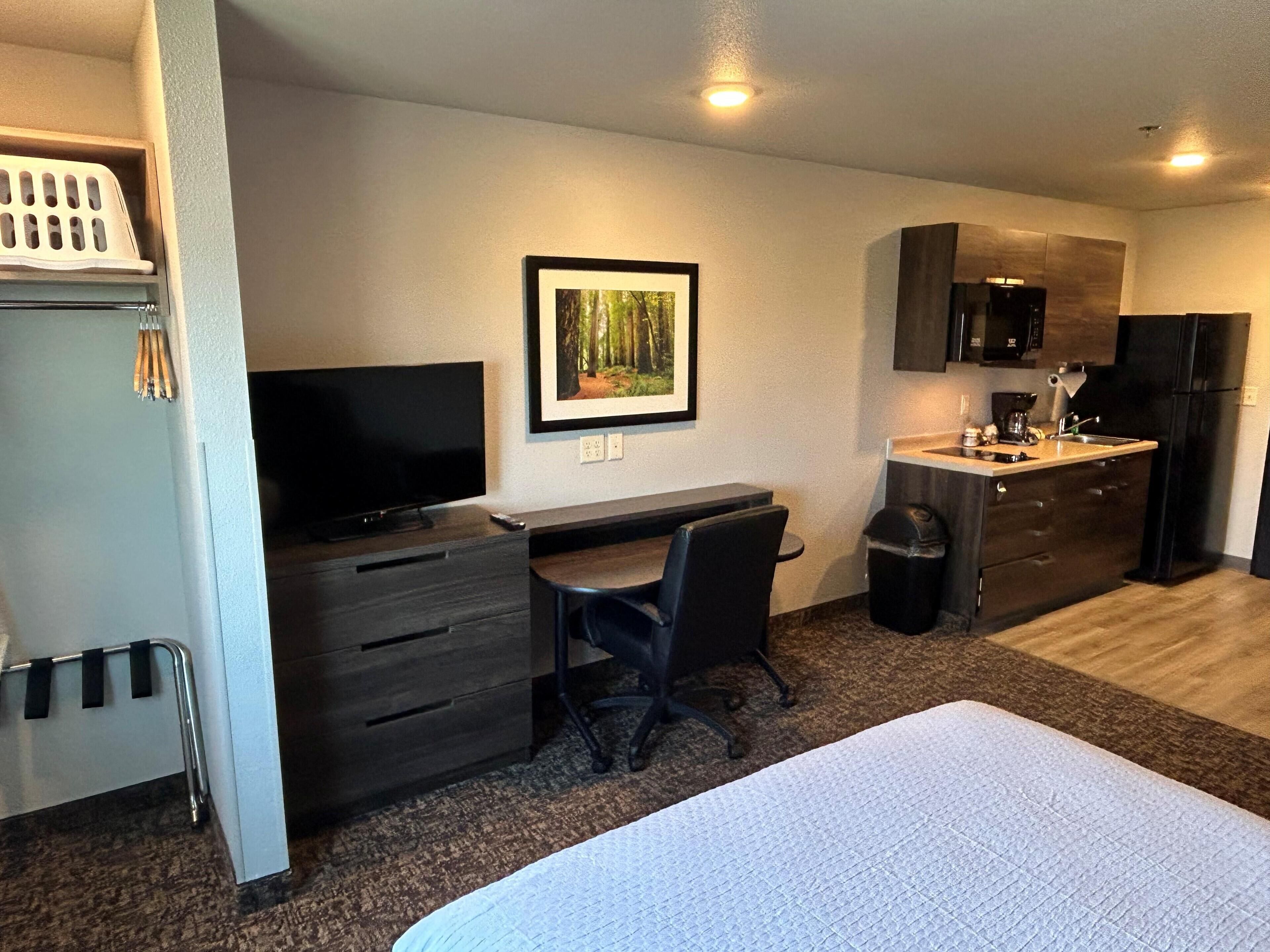 Una habitación de hotel compacta con cama, escritorio, silla, televisor, un cuadro enmarcado, una kitchenette pequeña y un cesto de basura.