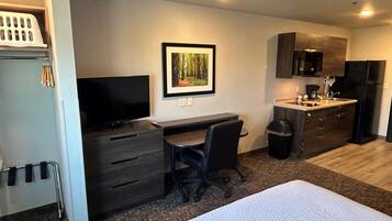 Una habitación de hotel compacta con cama, escritorio, silla, televisor, un cuadro enmarcado, una kitchenette pequeña y un cesto de basura.