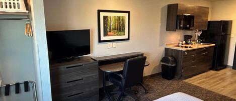A compact hotel room with a bed, a desk, a chair, a television, a framed picture, a small kitchenette, and a trash bin.