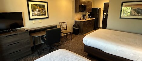 A hotel room with a bed, a desk with a chair, a television, a kitchenette, and framed pictures on the walls.
