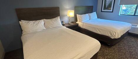Habitación de hotel con dos camas, una mesita de noche con lámpara, un cuadro enmarcado en la pared y una ventana con vistas.