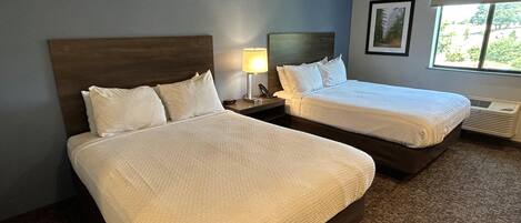 A hotel room with two beds, a nightstand with a lamp, a framed picture on the wall, and a window with a view of greenery.
