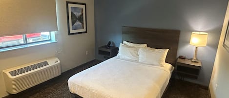 Une chambre d’hôtel avec un lit, une table de chevet, une lampe, un tableau au mur et une fenêtre avec des stores.