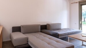 Un salon moderne avec un canapé d’angle, une petite table et une fenêtre équipée d’un appareil de climatisation.