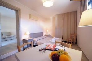 A hotel room with a bed, bedside table, lamp, and a dining table with a fruit bowl, glasses, and a magazine.