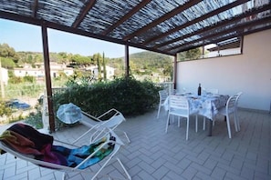 A covered patio with a table and chairs, a lounge chair, and a view of the surrounding landscape.