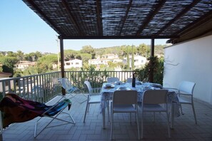 Uma área externa coberta com mesa e cadeiras, uma poltrona de descanso e vista para casas ao fundo.