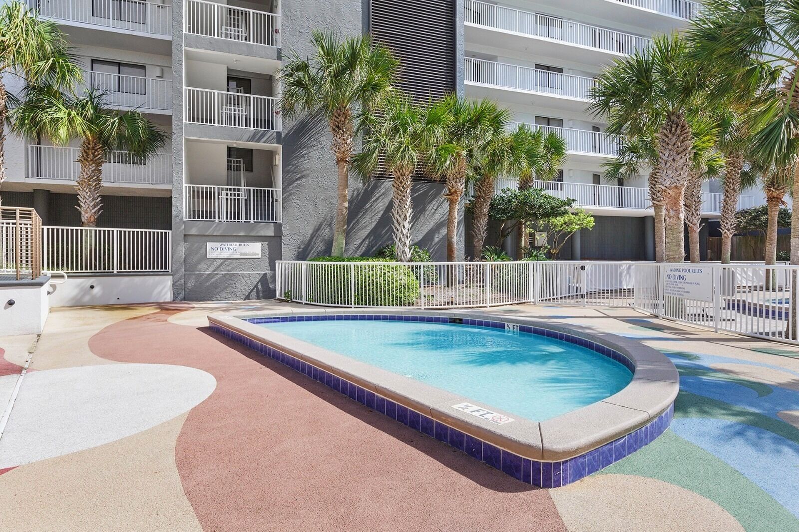 A swimming pool surrounded by a white fence, with palm trees and a multi-story building in the background.