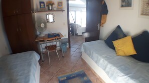 Une petite pièce bien éclairée, avec deux lits, un bureau, une chaise et un ventilateur.