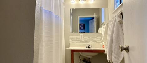A bathroom with a shower curtain, a sink, a mirror, and a window with a bottle of air freshener.