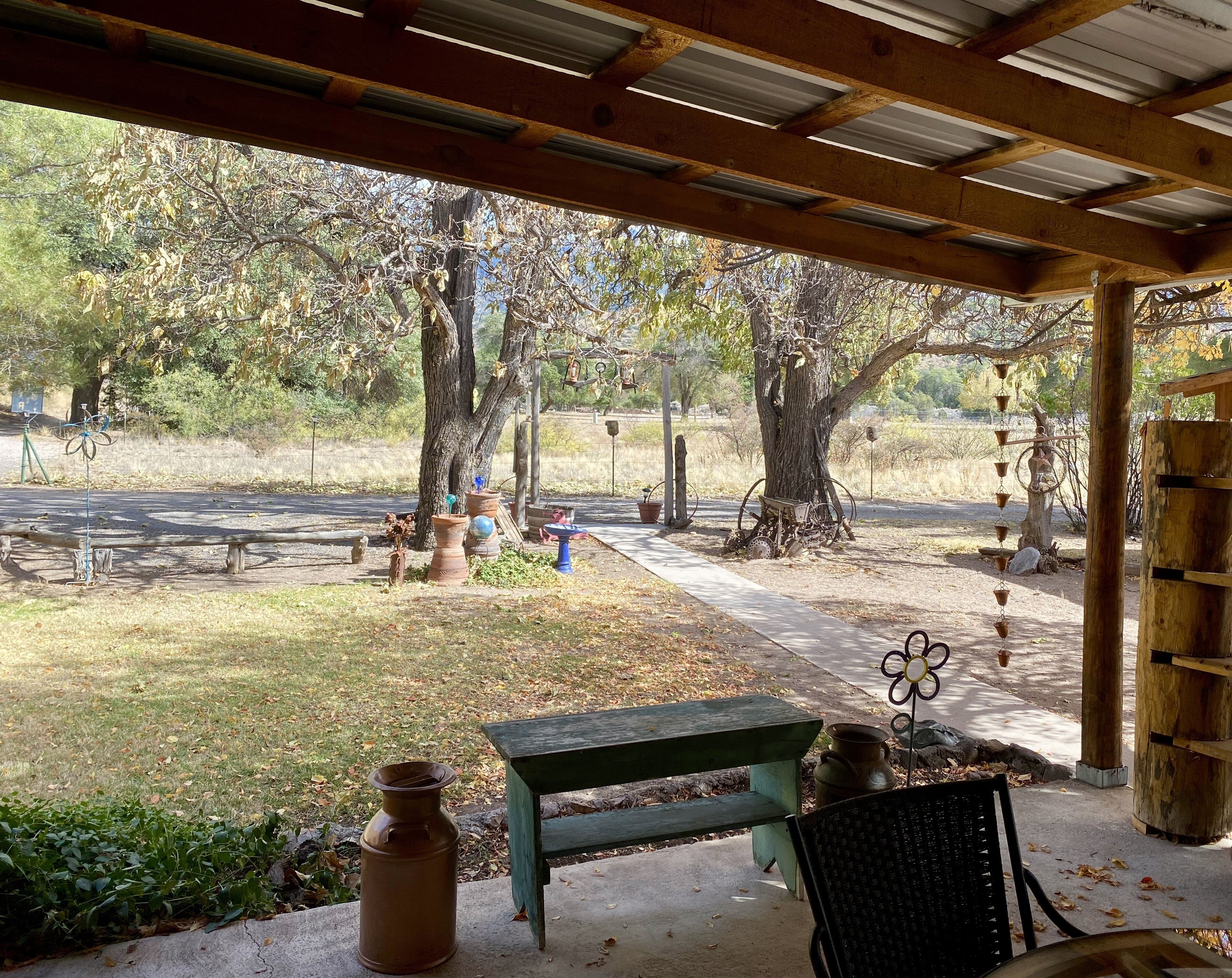 A covered patio with a green bench and a table, surrounded by trees and a paved path.