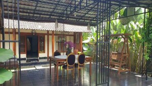 A covered patio area with a dining table and chairs, surrounded by plants and a wooden swing. - OYO 3211 Ndalem Dasuriah (Minggir)