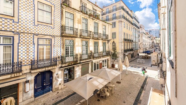 A narrow street with buildings featuring traditional architecture, a cobblestone ground, and outdoor seating areas. - GuestReady - Apt located on Lisbon city centre (Lisbon)
