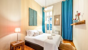 A bedroom with a bed, a bedside table, a lamp, a window with curtains, a radiator, and a shelf with flowers. - GuestReady - Apt located on Lisbon city centre (Lisbon)