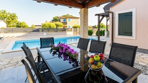 Ein überdachter Sitzbereich im Freien mit Pool, Tisch mit Stühlen und einer Obstschale.