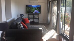 Ein Wohnzimmer mit einer Couch, einem Fernseher und Blick auf einen Garten durch große Fenster.