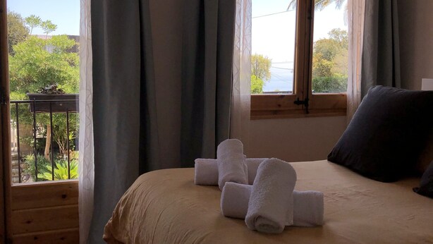 Ein Schlafzimmer mit Bett, Kissen und Handtüchern. Es gibt Fenster mit Vorhängen, einen Balkon und Blick ins Grüne.