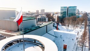 Un bâtiment moderne, orné d’un mât sur lequel flotte un drapeau, avec un sol recouvert de neige et une vue dégagée sur les contours de la ville en arrière-plan.