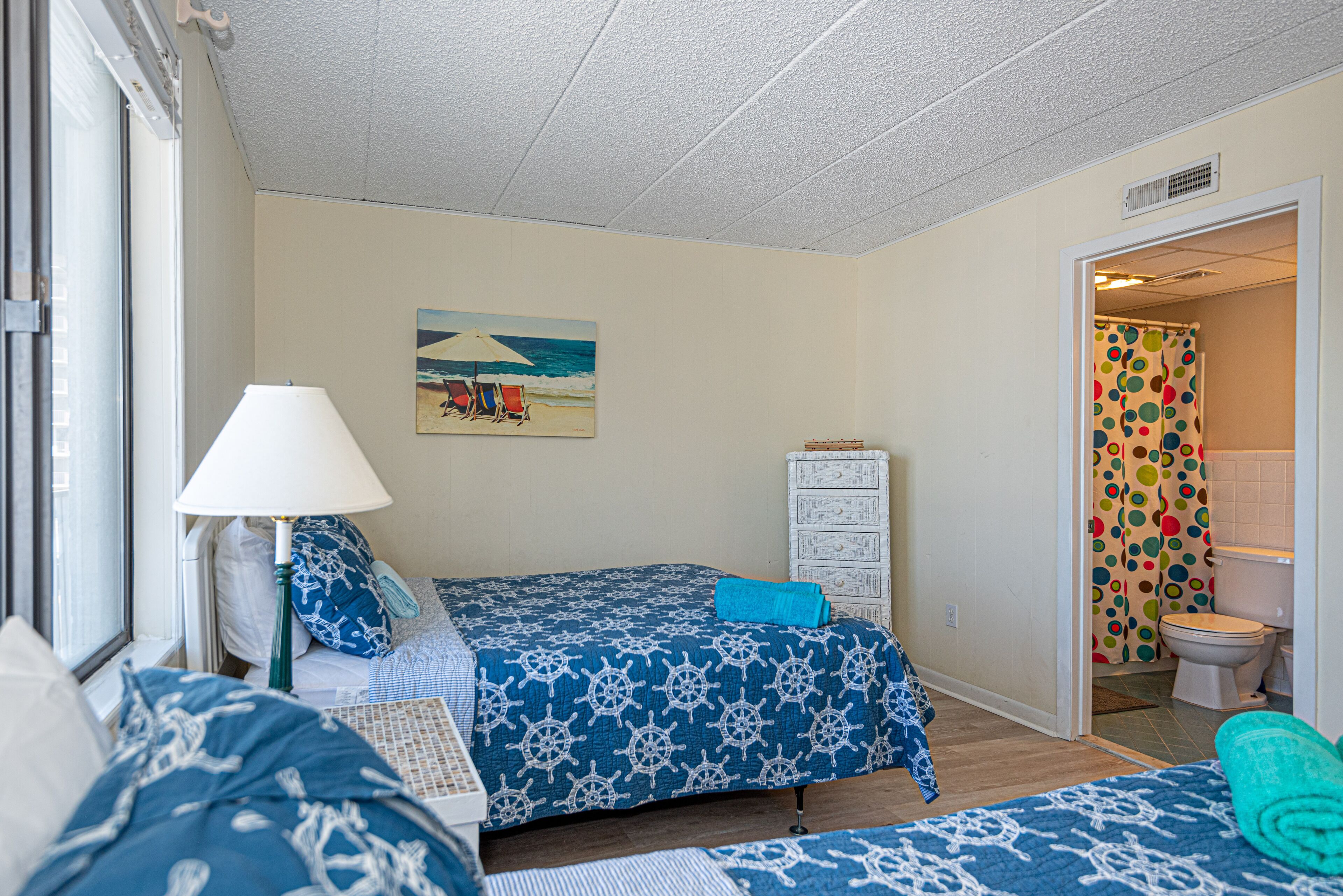 A bedroom with a bed, a nightstand, a lamp, a dresser, and a painting of a beach scene.