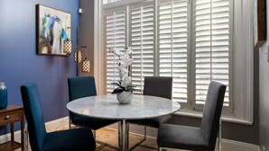 A dining area with a round table, four chairs, a vase with flowers, a painting on the wall, and a window with blinds.