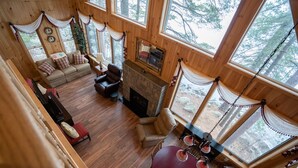 Interior acogedor de una cabaña con chimenea, pisos de madera, un sofá y grandes ventanas que ofrecen vistas del bosque circundante.