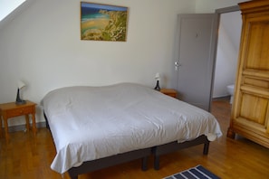A bedroom with a large bed, a nightstand with a lamp, a wooden wardrobe, and a door leading to another room.