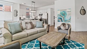 Ein modernes Wohnzimmer mit einer Couch, einem Couchtisch und einem Sessel. Im Hintergrund befindet sich eine Küche mit weißen Schränken und einem Essbereich.