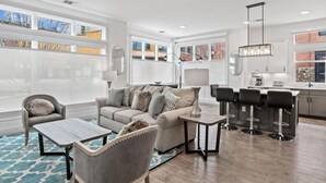 Ein geräumiges Wohnzimmer mit einer Couch, Sesseln und einem Couchtisch. Es gibt eine Küchenzeile mit Barhocker und einer Pendelleuchte.