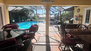 An outdoor pool area with lounge chairs and a dining table, surrounded by potted plants and a glass enclosure. - Gorgeous waterfront Spanish villa with heated pool on beautiful oversized canal! (Cape Coral)
