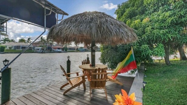 A wooden deck with a thatched umbrella, a flag, and a table with chairs. - Gorgeous waterfront Spanish villa with heated pool on beautiful oversized canal! (Cape Coral)