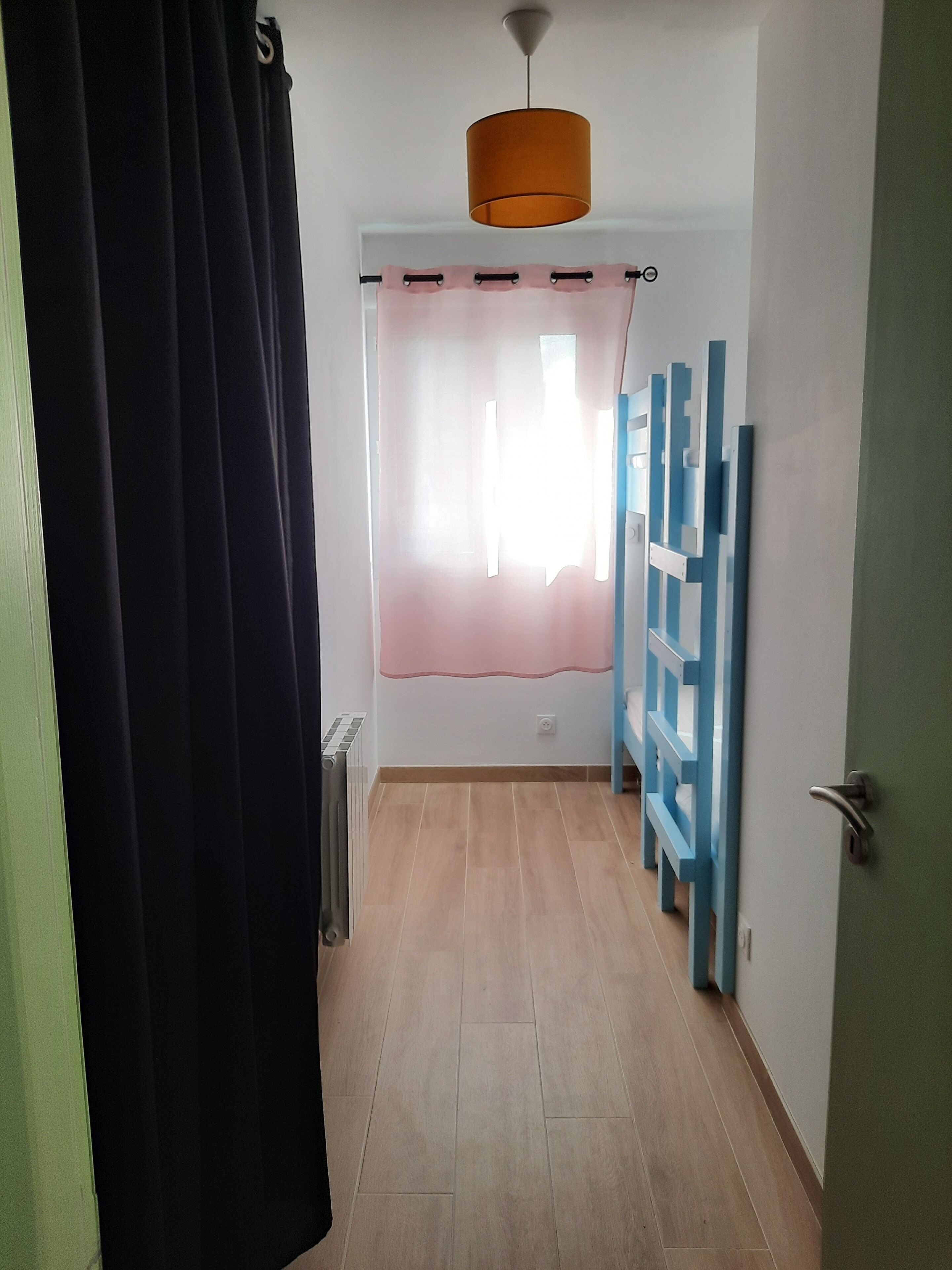 Un pasillo con un radiador, una lámpara en el techo, una ventana con cortina rosa y un marco de puerta azul.