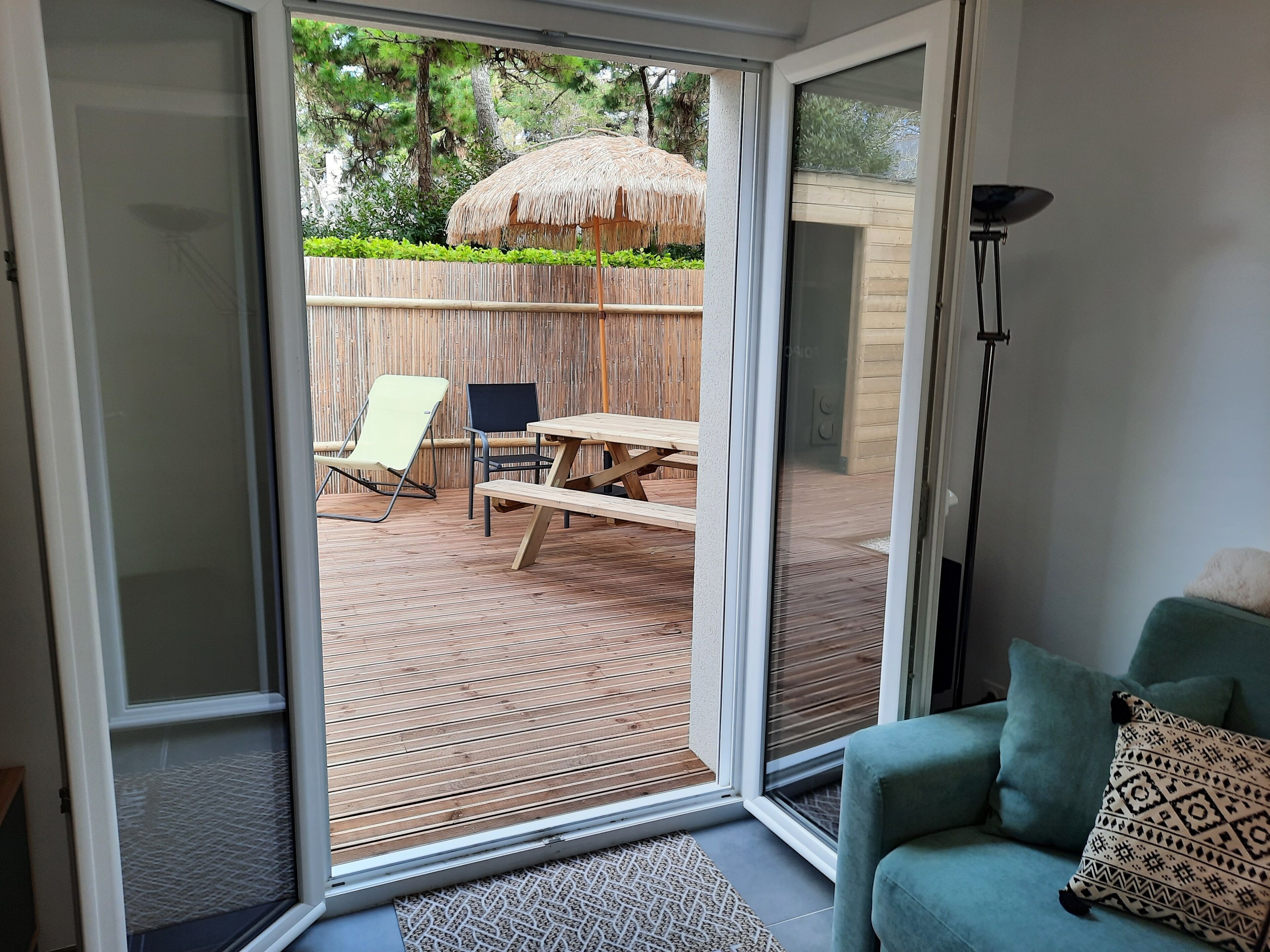 Eine Glastür zur Terrasse mit Holzdeck, einem Reetdachschirm und einem Picknicktisch.