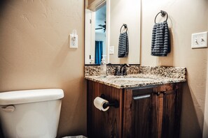 Ein Badezimmer mit einem Holzbadezimmerhocker, einem Waschbecken mit geflecktem Waschtisch, einer Toilette und einem Spiegel über dem Badehocker.
