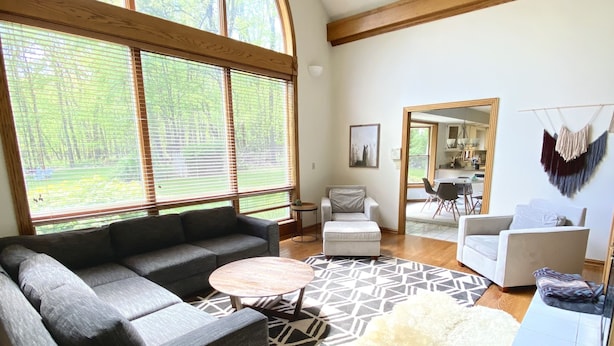 A spacious living room with a large window, a grey sectional sofa, a round coffee table, and a white shag rug. - 30 min from Notre Dame + Hottub, 10 acre estate and game room (Edwardsburg)