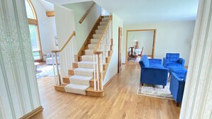 A spacious living area with a wooden staircase, blue furniture, and a dining area in the background. - 30 min from Notre Dame + Hottub, 10 acre estate and game room (Edwardsburg)