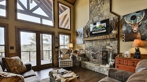 A spacious living room with a stone fireplace, large windows, and rustic decor. - Mountain River Lodge by Mountain Oasis | River View Cabin in Ellijay (Ellijay)