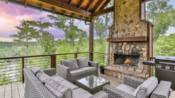 An outdoor deck with wicker furniture, a stone fireplace, and a grill. - Mountain River Lodge by Mountain Oasis | River View Cabin in Ellijay (Ellijay)