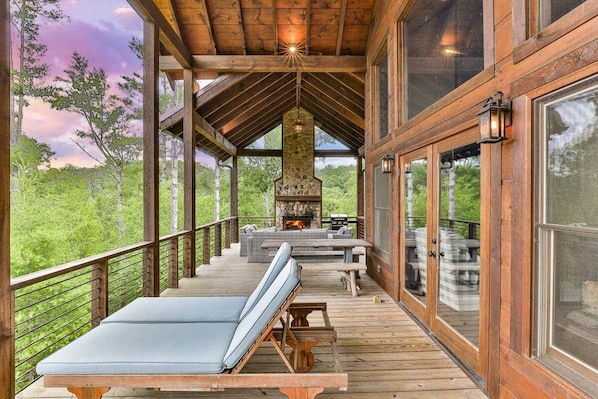 A wooden deck with a stone fireplace, outdoor seating, and a view of the forest.