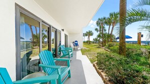 A covered outdoor seating area with turquoise chairs and a table, overlooking a beach with palm trees and a pier. - Ground-Floor | Huge Patio | Steps to Pool and Beach | FLPCB9 (Panama City Beach)