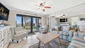 A spacious living room with a ceiling fan, a sofa, a coffee table, and a television. - Ground-Floor | Huge Patio | Steps to Pool and Beach | FLPCB9 (Panama City Beach)