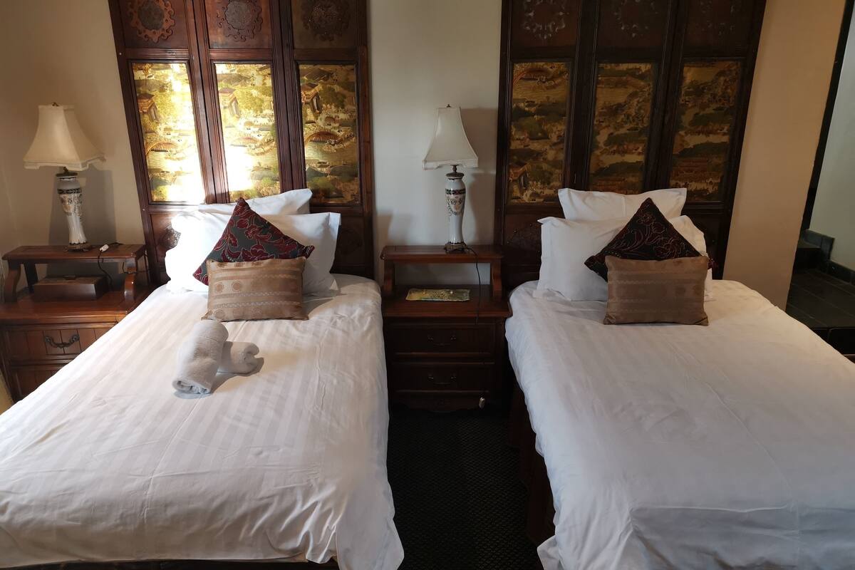 Een hotelkamer met twee eenpersoonsbedden, elk voorzien van witte lakens en decoratieve kussens. Twee nachtkastjes met lampen en een kleine tafel tussen de bedden.