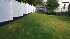 Een verzorgd grasveld met een witte muur en een metalen poort, leidend naar een gebouw met een overdekt terras.