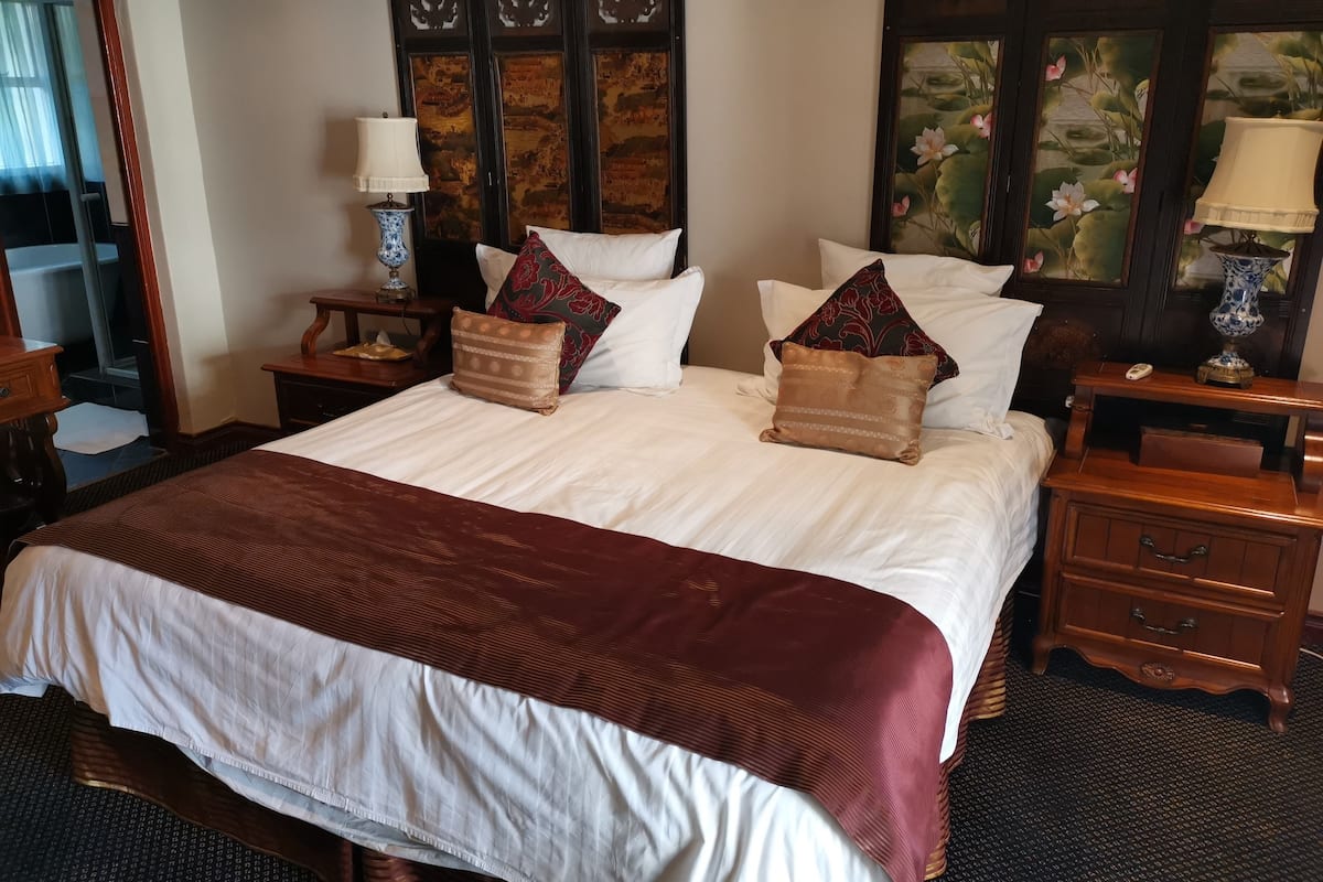 Een net opgemaakt bed met een bruine dekbedovertrek en meerdere kussens, een houten nachtkastje met een lamp, en een wand met een decoratief paneel.