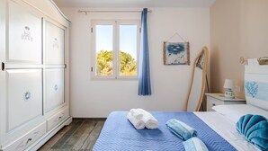 A bedroom with a bed, a wardrobe, a window with curtains, a bedside table with a lamp, and a wall decoration. - Vacation home with sea view Ogliastra Sardinia Casa Lentisco D (Foxi Murdegu)