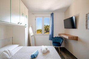 Habitación de hotel con cama, una silla, televisor, ventana con cortinas y repisa en la pared.