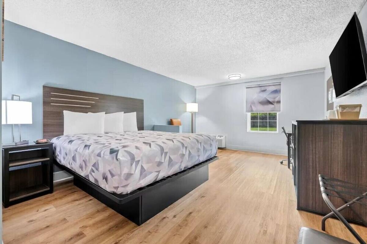 A modern hotel room with a bed, bedside tables, a desk, a chair, a flat-screen TV, and a window.