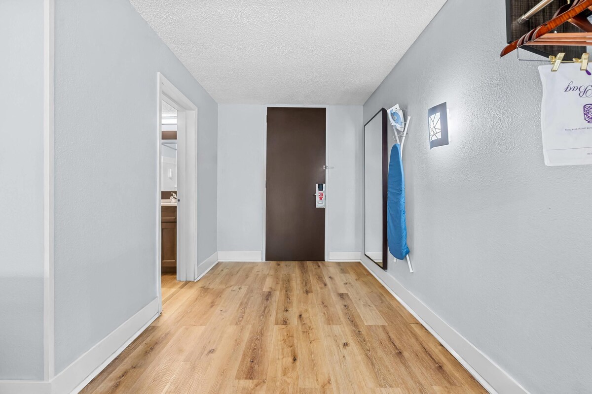 A hallway with wooden flooring, light gray walls, and a door leading to a room.
